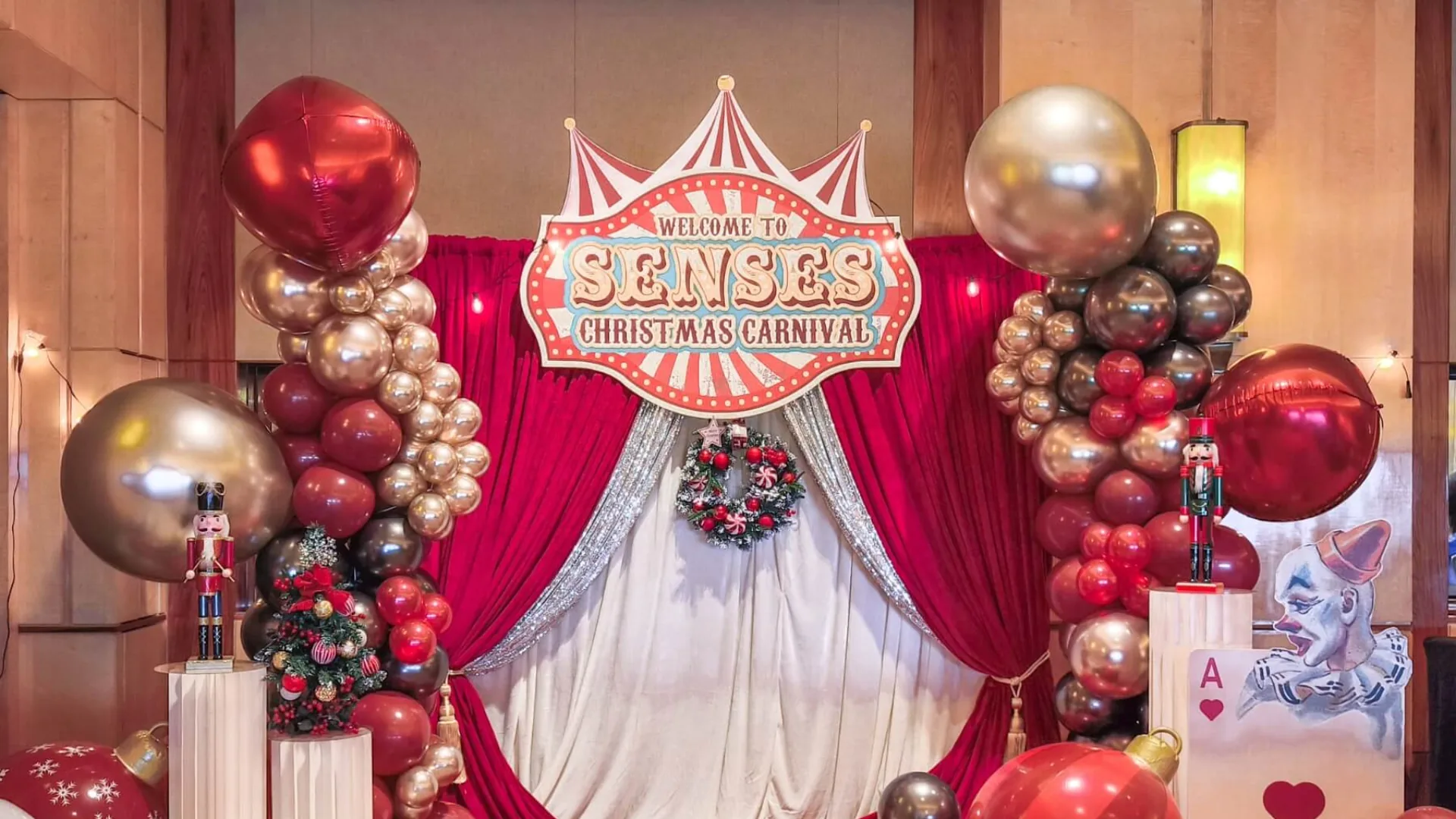 Senses Christmas Carnival photo backdrop with a big-top welcome sign, red drapes and red and gold balloon columns