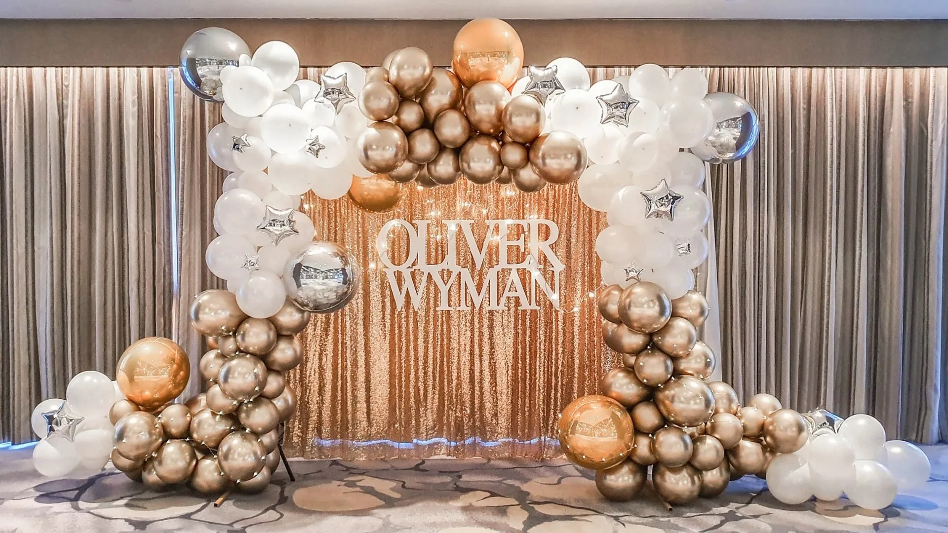 Elegant Oliver Wyman company celebration backdrop with white, gold and bronze balloon styling